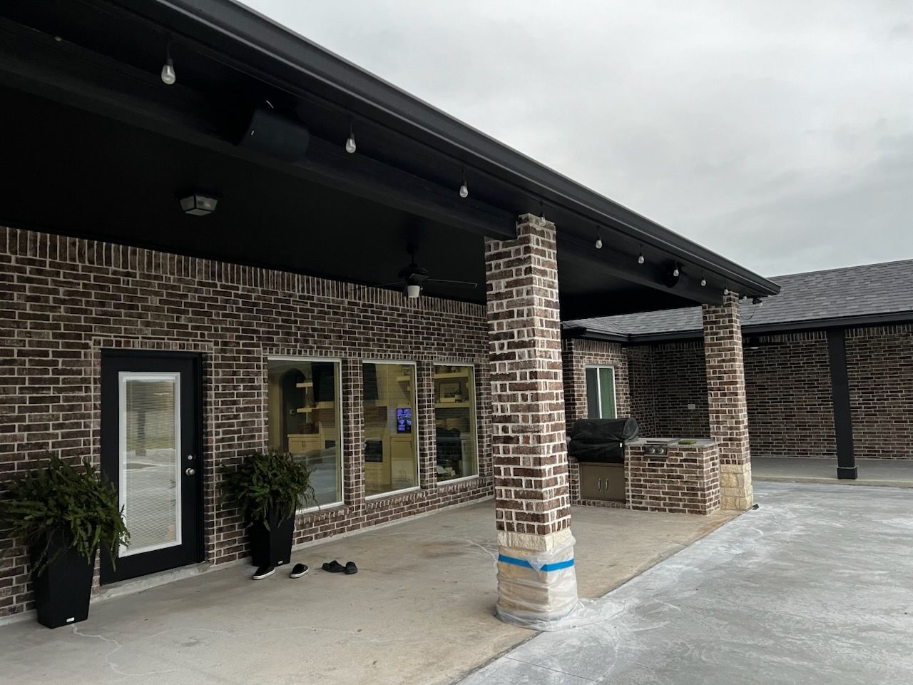 A covered brick patio features a doorway, large windows, an outdoor grill area, and potted plants under a black ceiling.