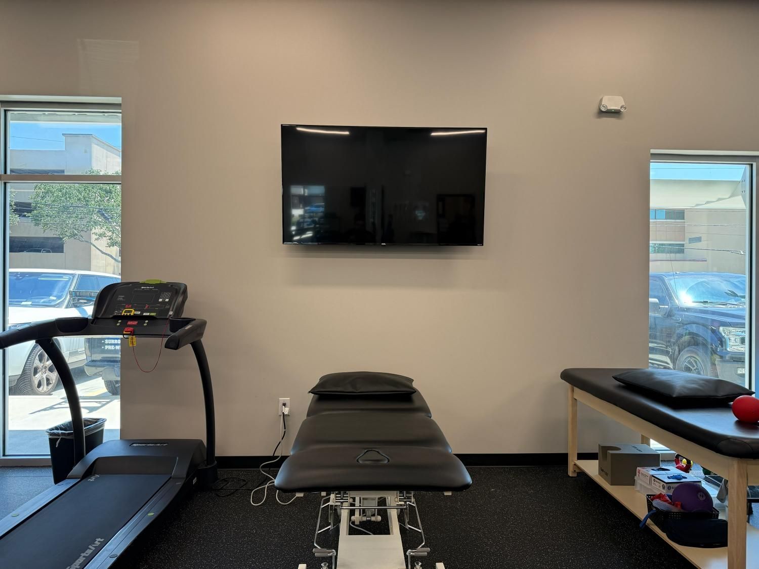 Physical therapy room with a treadmill, treatment tables, and a wall-mounted TV.