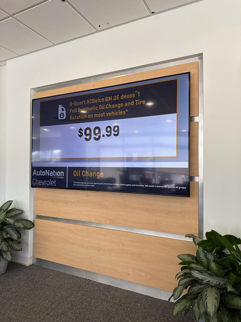 Digital sign displaying $99.99 price, possibly for EV charging, mounted on wooden panel with plants.
