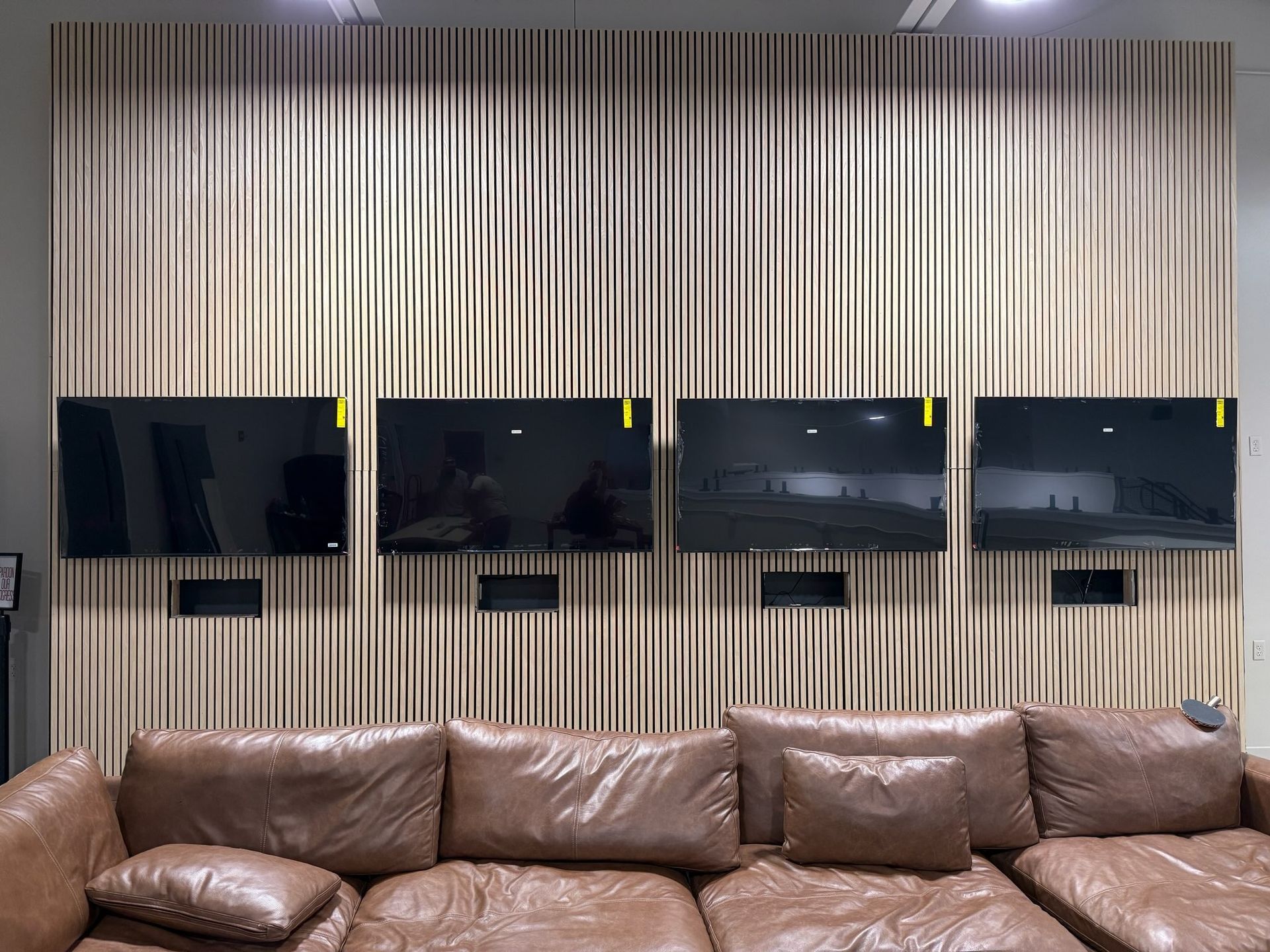 Four TVs mounted on a wood-paneled wall above a brown leather couch.