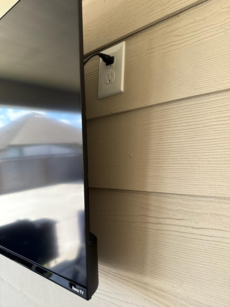 Black TV mounted on a tan siding wall, with power cord plugged into an outlet.