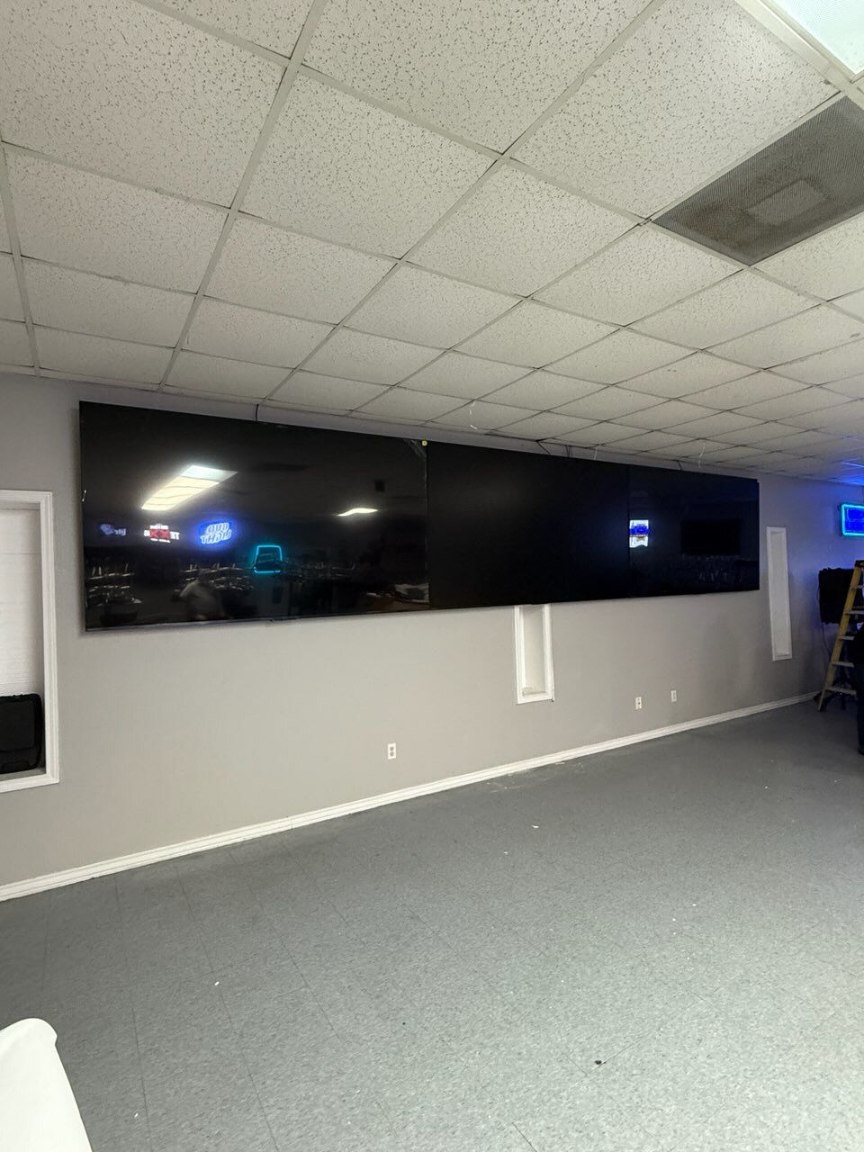 Large black TV mounted on a gray wall in a room with a drop ceiling and gray carpet.