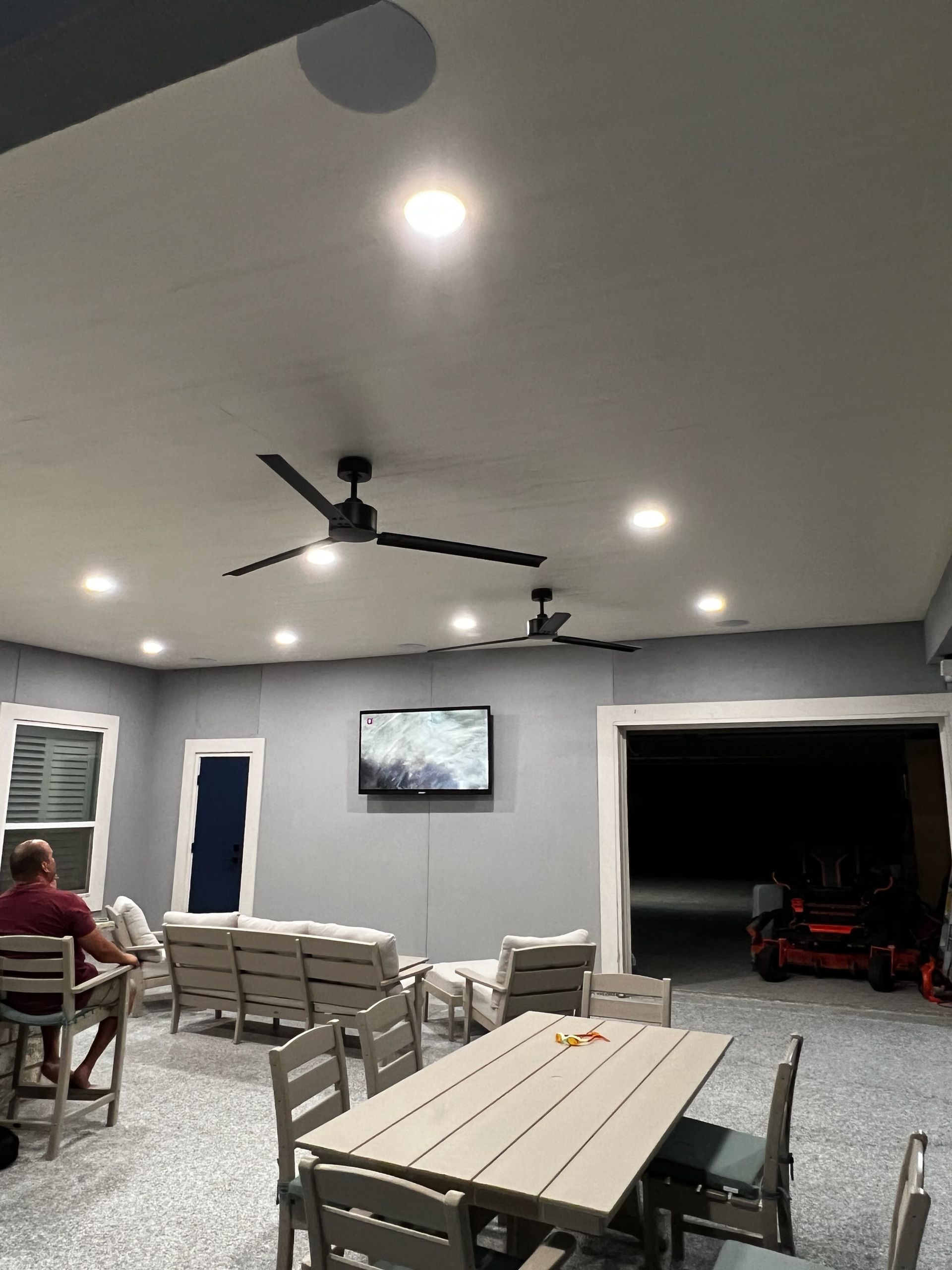 An outdoor covered patio features a dining table with chairs, a sectional sofa, a wall-mounted TV, and ceiling fans.