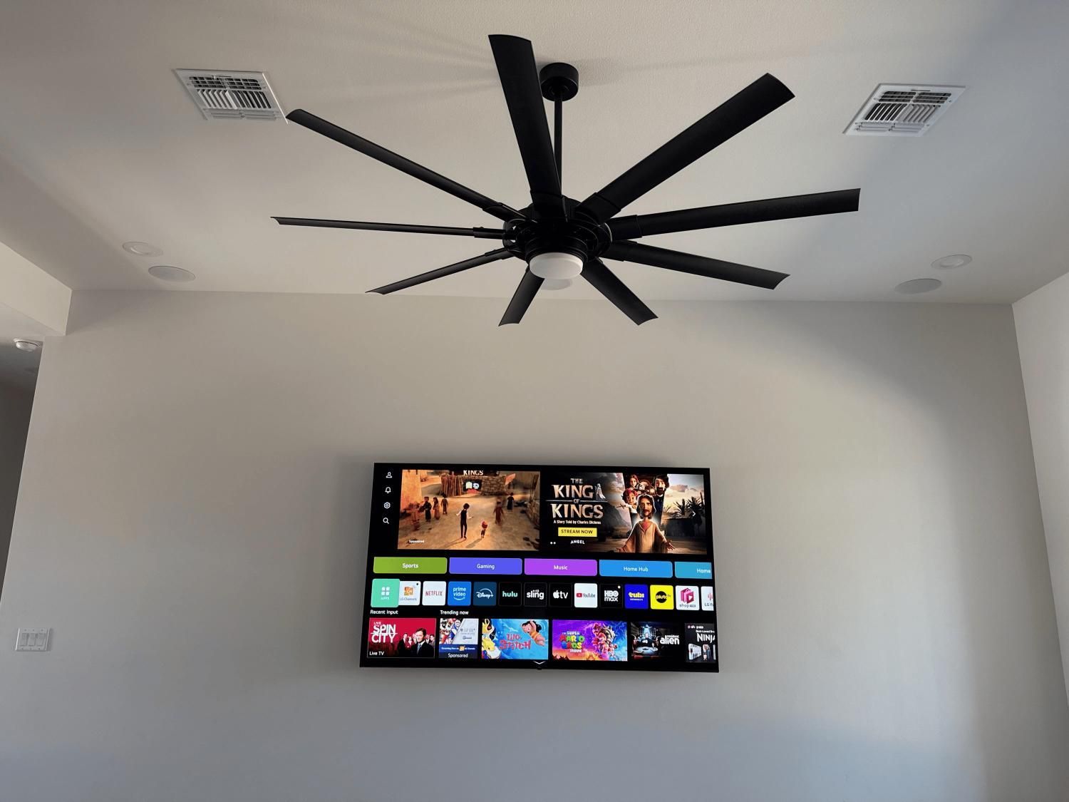 Black ceiling fan above a mounted TV. White walls, air vents visible. TV shows a streaming app interface.