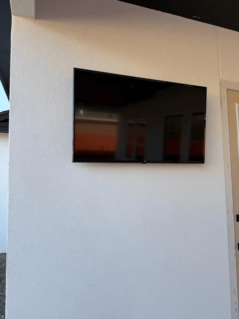 Black television mounted on a textured white stucco wall outdoors, reflecting the surroundings.