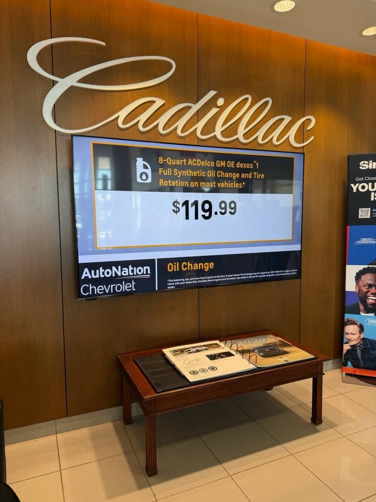 Cadillac dealership interior with a digital display showing a service special for $119.99, mounted on a wood-paneled wall.
