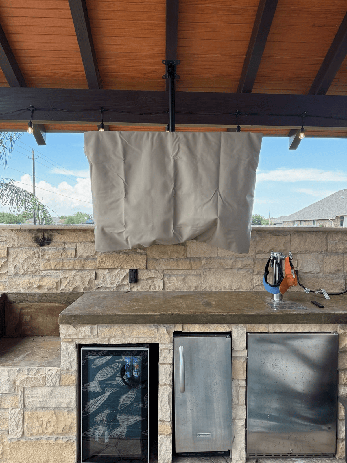 Outdoor kitchen with a covered TV, stone counter and cabinets, and a blue sky.