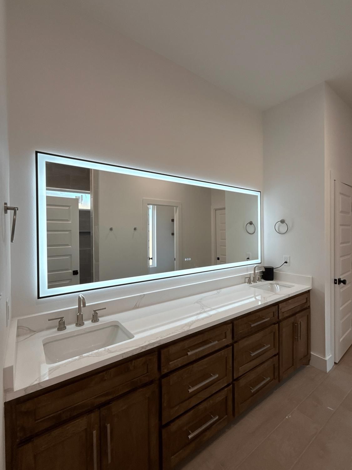 Modern bathroom with large backlit mirror over a long wooden vanity with white countertop and sink.