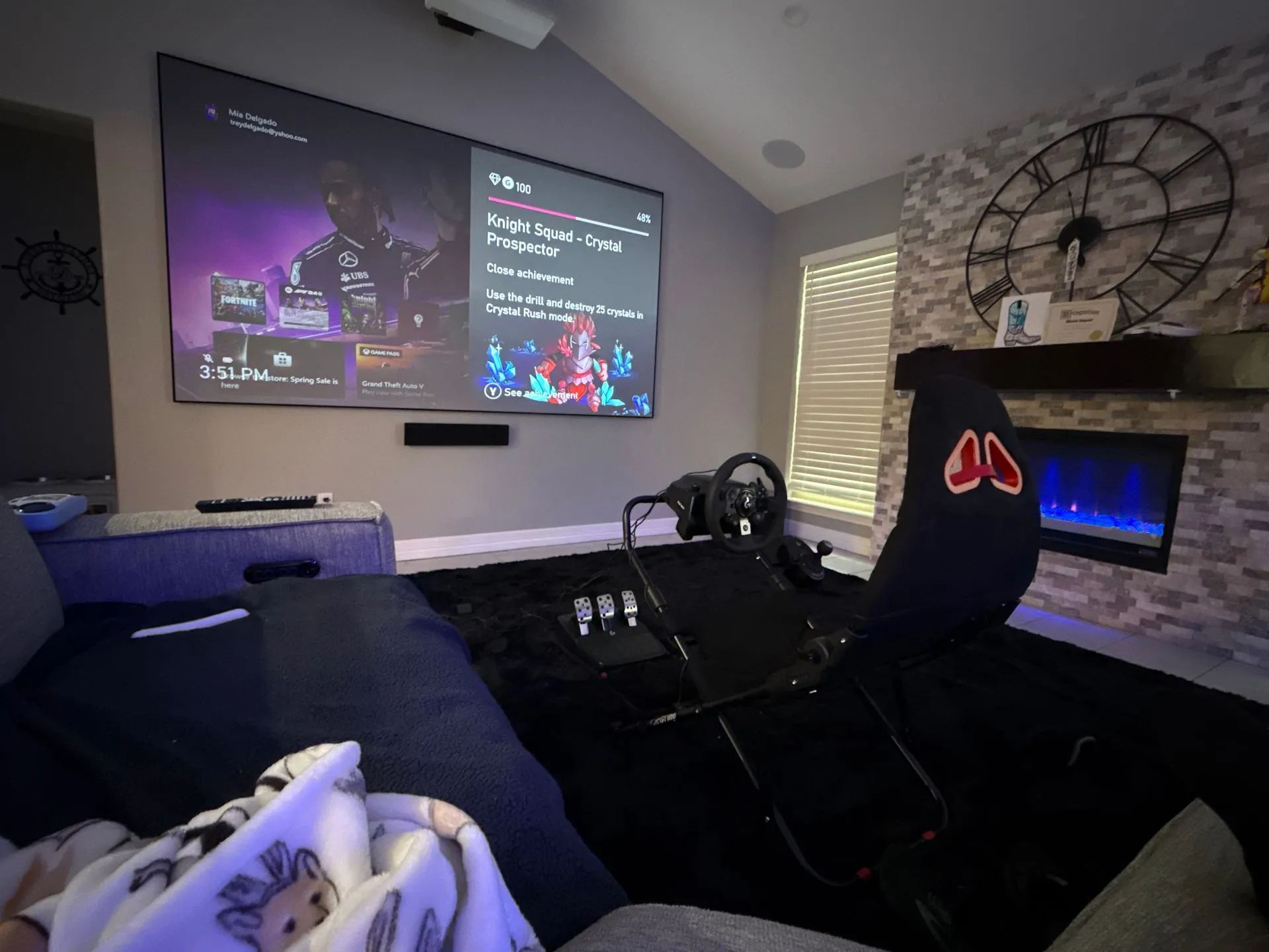 A home entertainment setup features a racing simulator chair in front of a large screen displaying video game content.