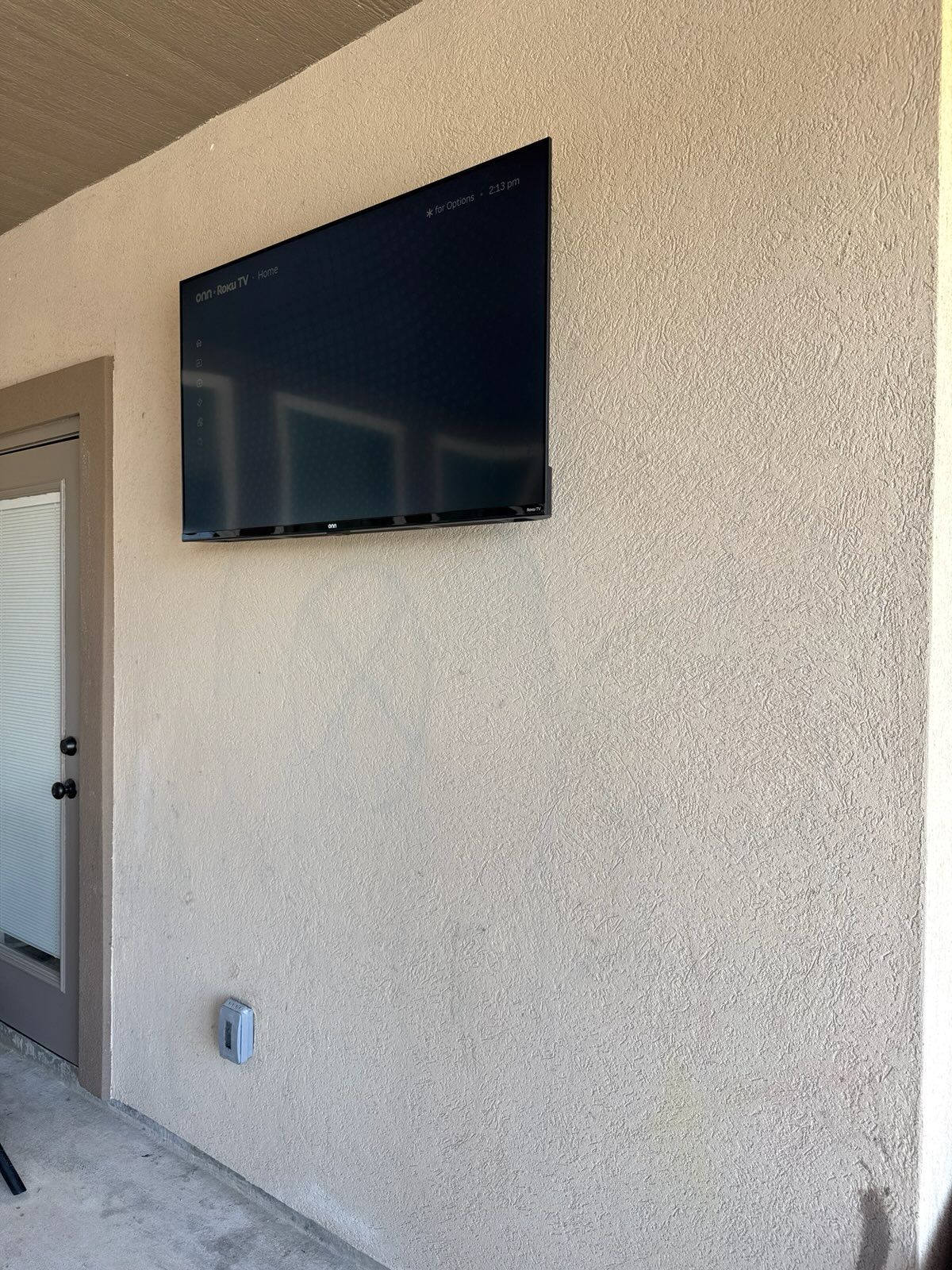 Wall-mounted TV on a beige stucco patio wall beside a door and outdoor light fixture.