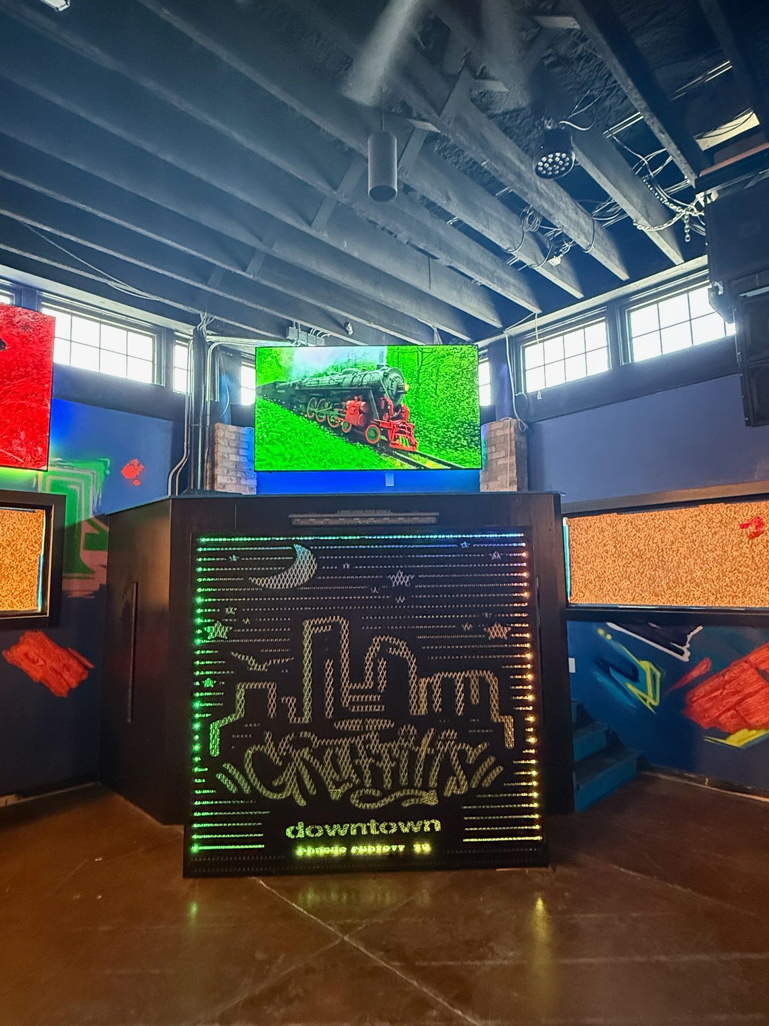 Indoor concert stage with large LED screens, colorful abstract visuals, and a central DJ booth under exposed rafters