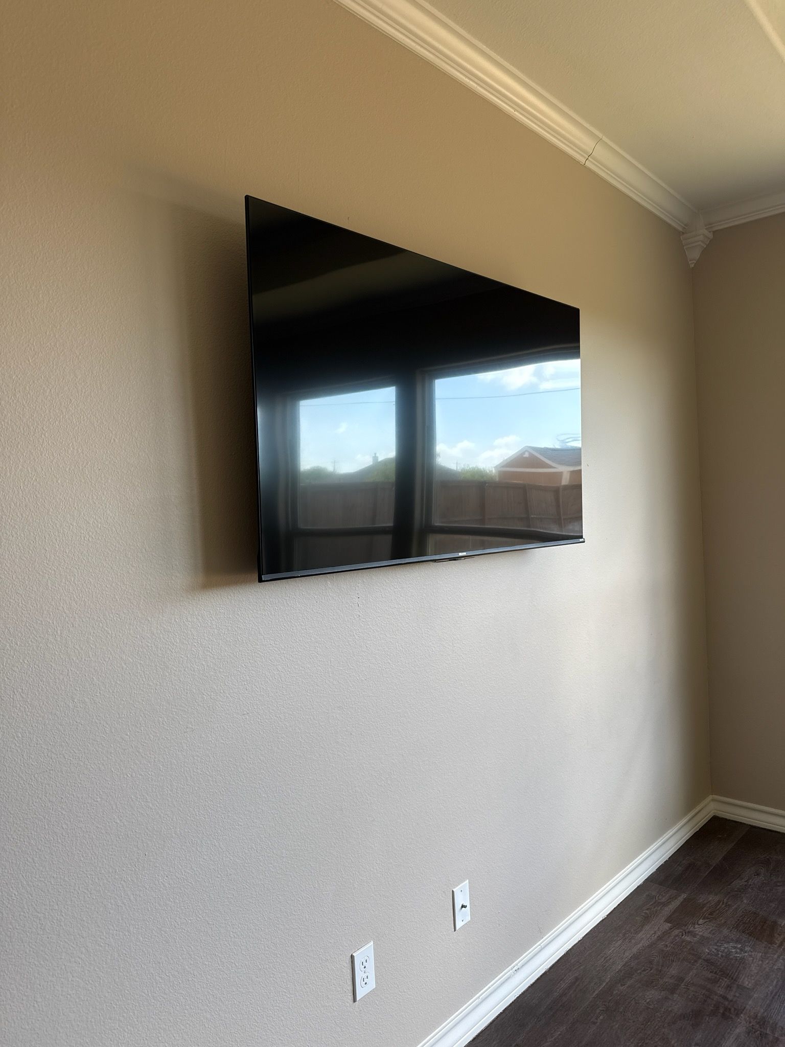 Wall-mounted flat-screen TV on a beige living room wall with dark carpet and windows reflected on the screen.