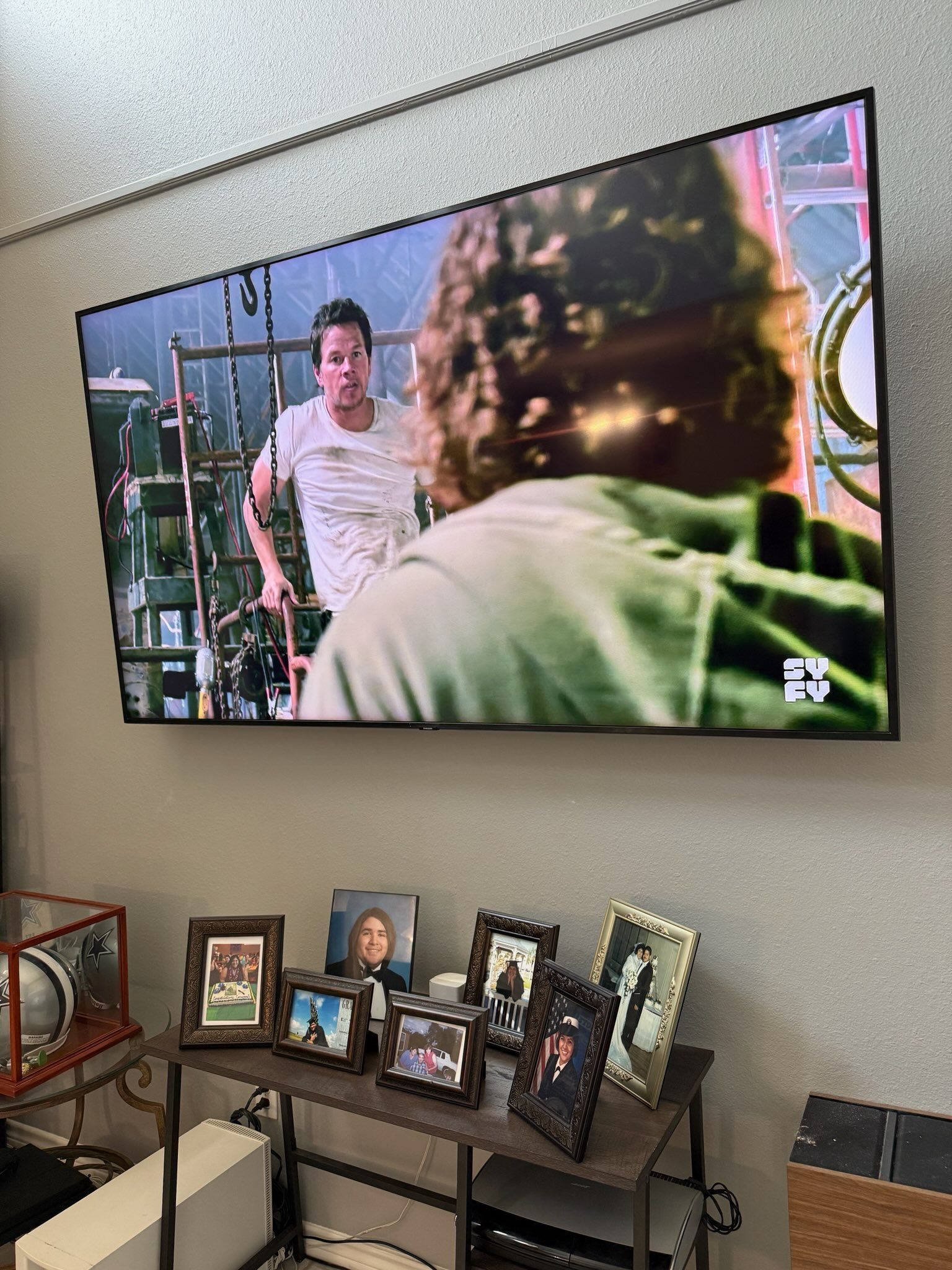 Wall-mounted TV showing two people indoors above a console with framed family photos