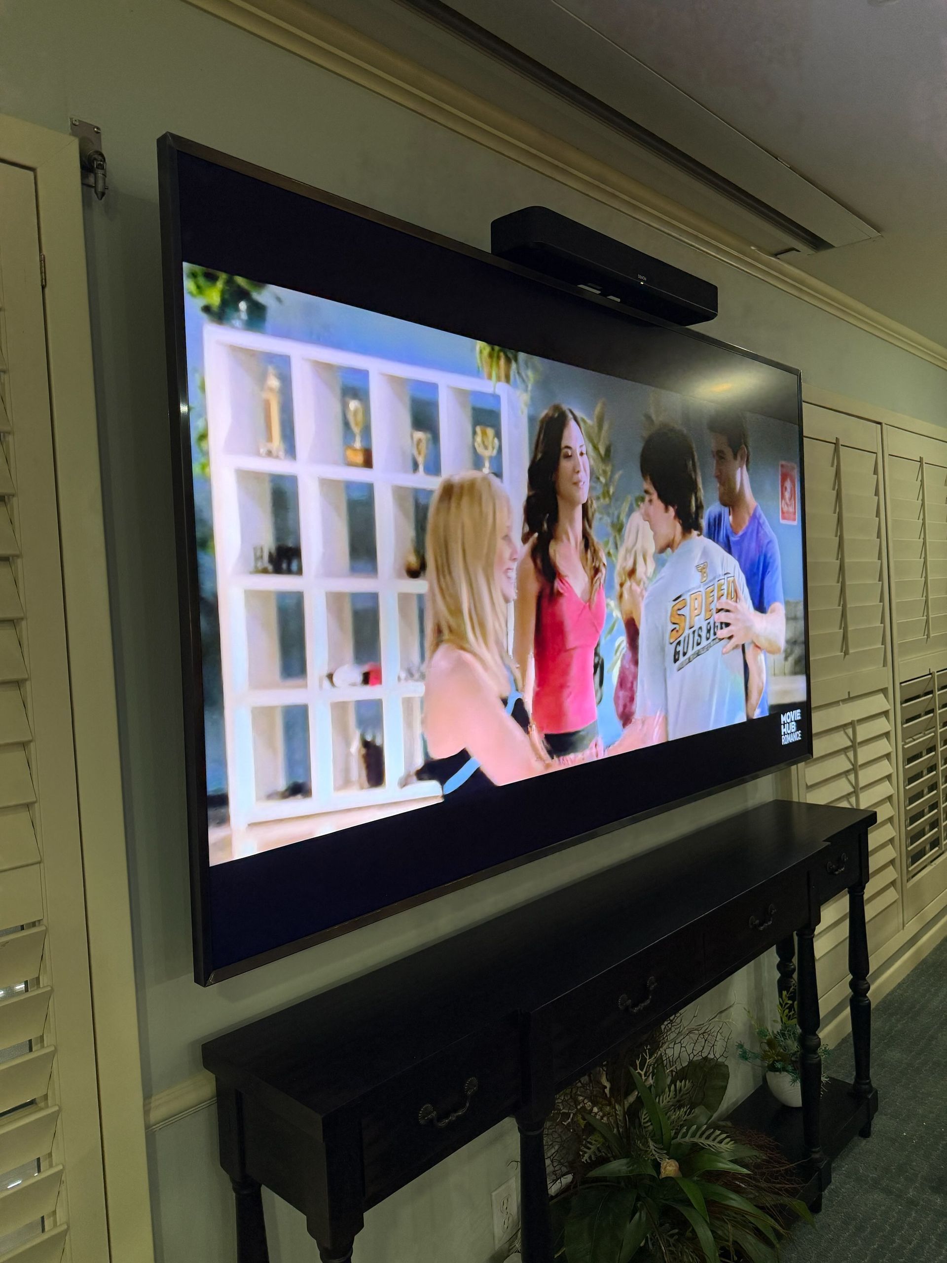 Wall-mounted TV shows three people talking in a bright room, with a black console beneath it.