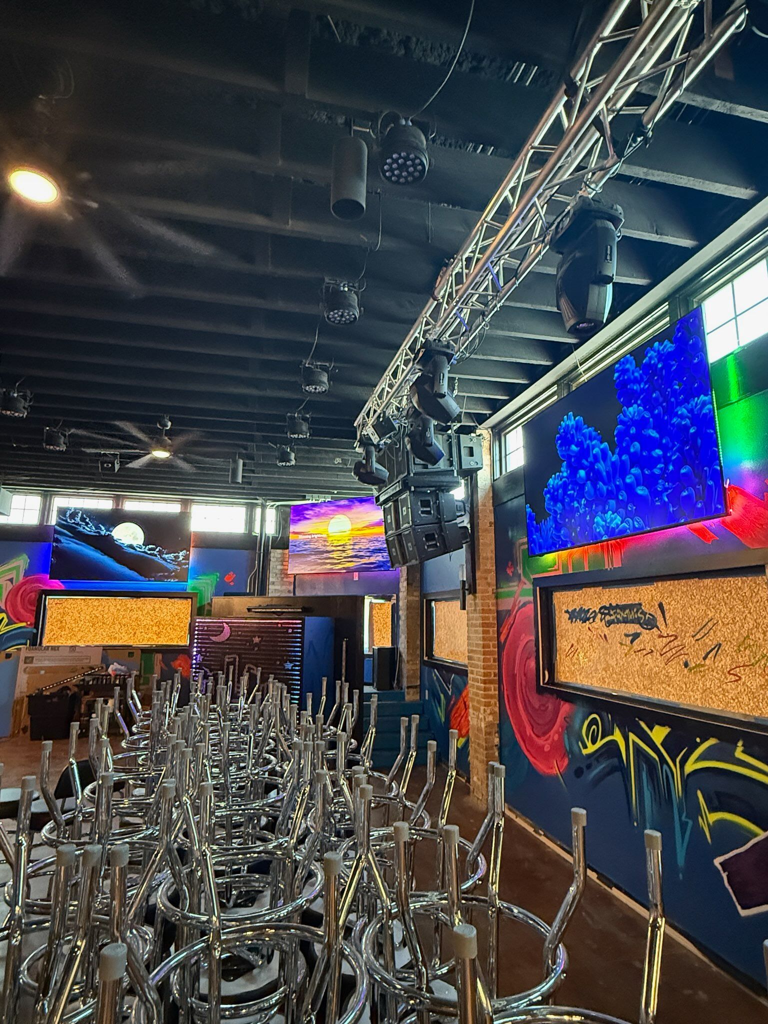 Empty nightclub with stacked chairs, neon wall art, and colorful screens under a dark ceiling.