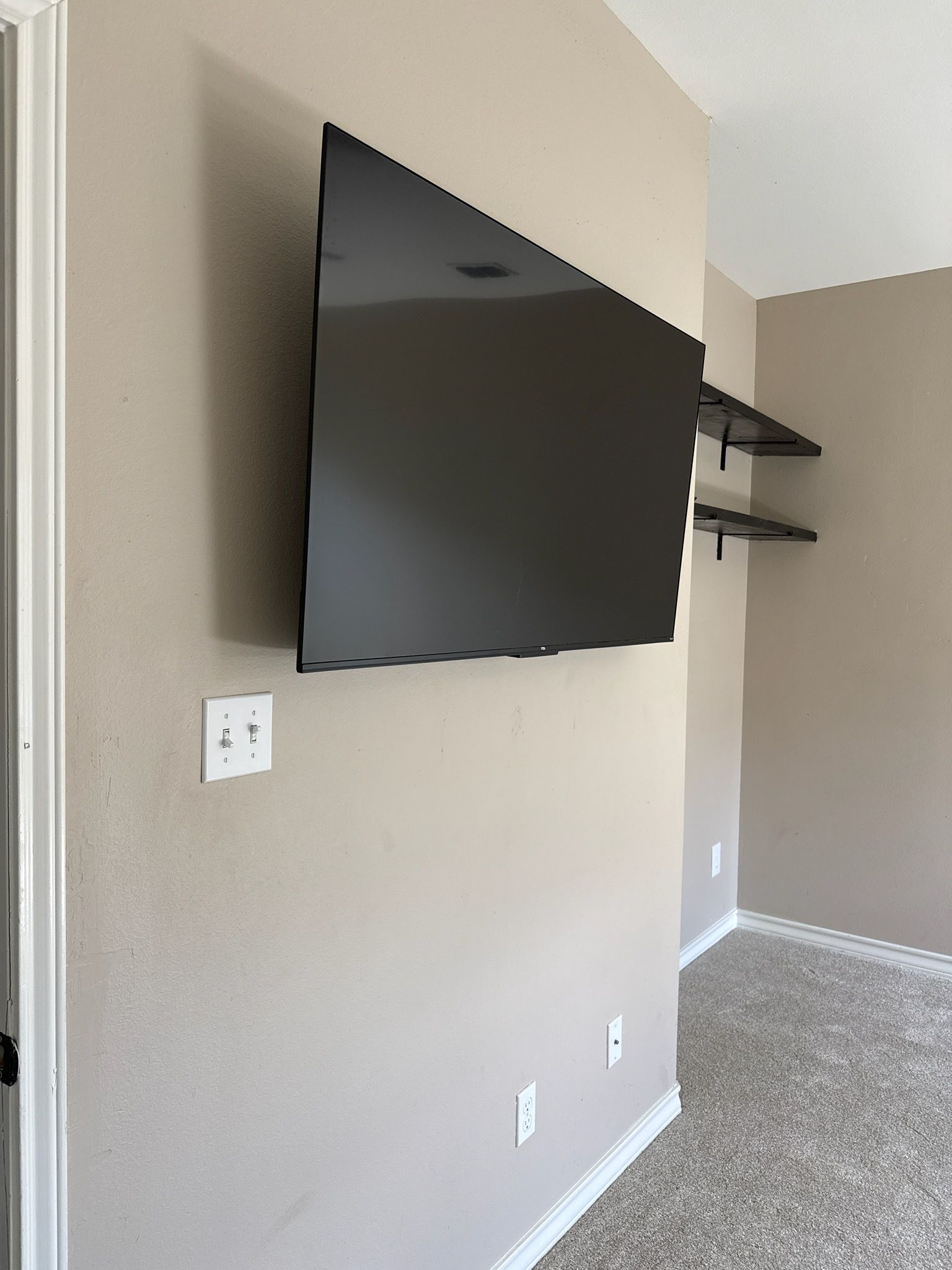 Living room wall with mounted flat-screen TV, floating shelves, and beige carpet.