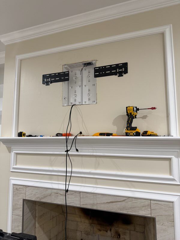 Wall-mounted TV bracket with exposed cables above a white fireplace mantel; drill and tools on the mantel.