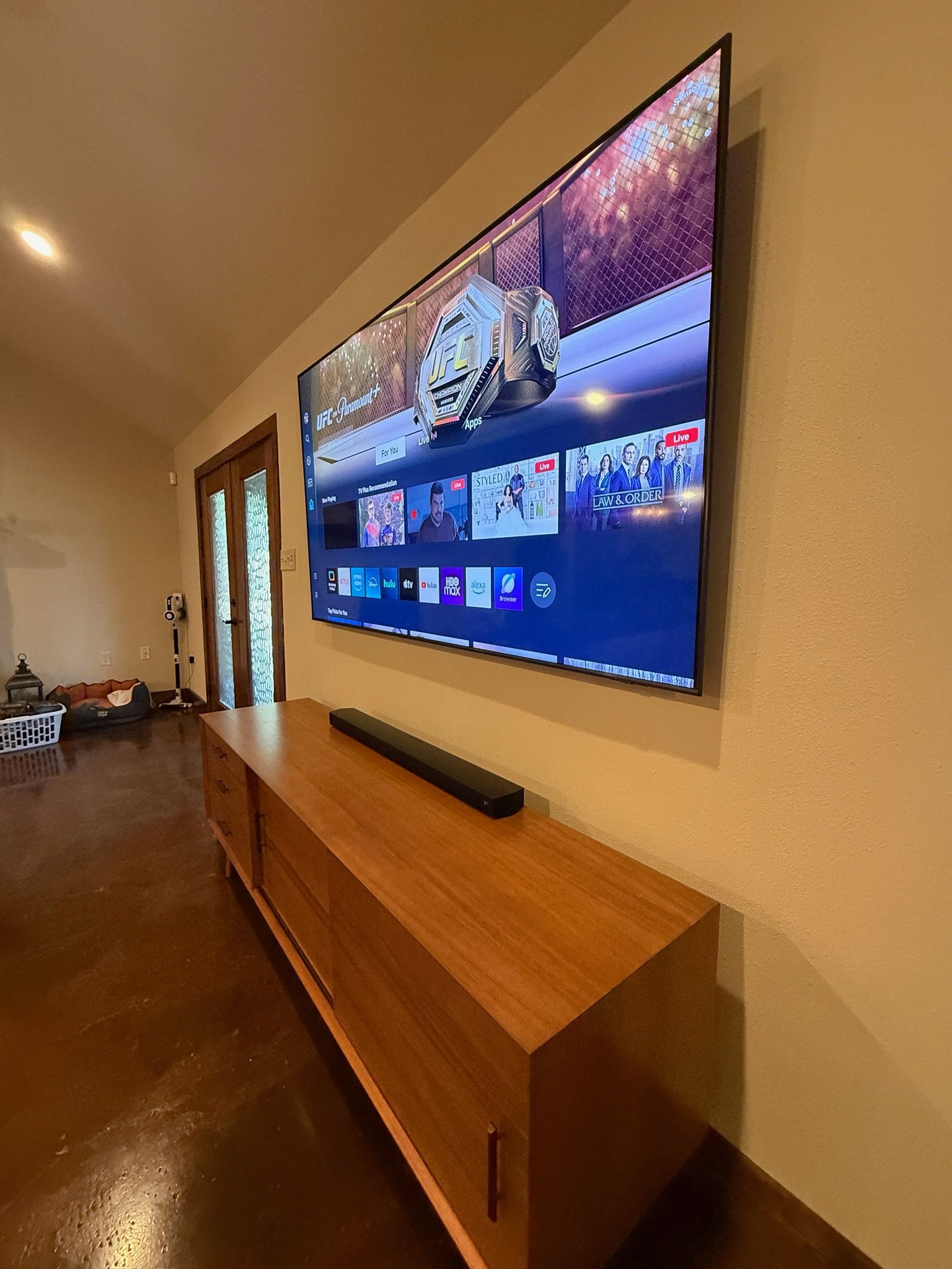 Wall-mounted TV in a hallway above a wooden console with a soundbar, showing a streaming menu.