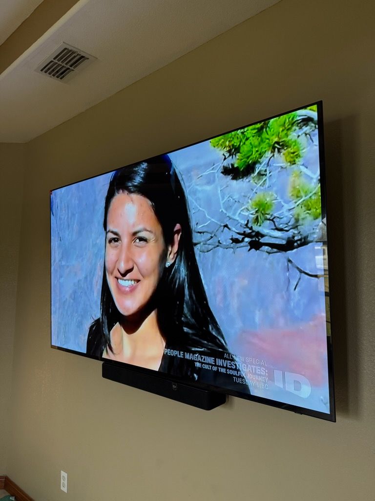 Wall-mounted TV shows a smiling woman beside water and green trees in a room with beige walls.