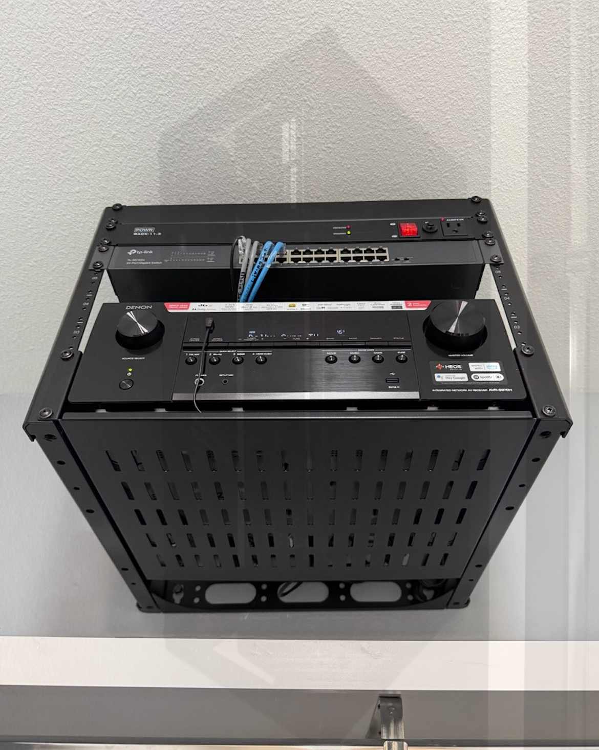 Black AV equipment rack mounted on a white wall, holding a receiver, network switch, and other electronics.