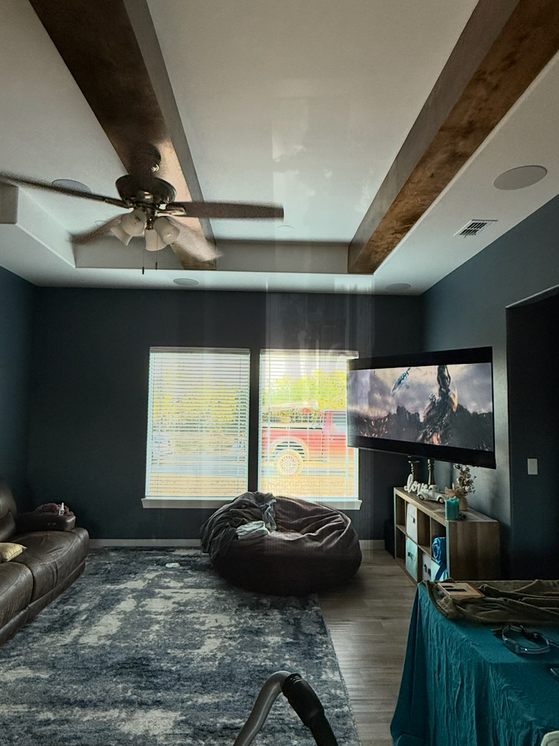 Living room with dark blue walls, light ceiling with wood beams, rug, TV, beanbag chair, and two windows.
