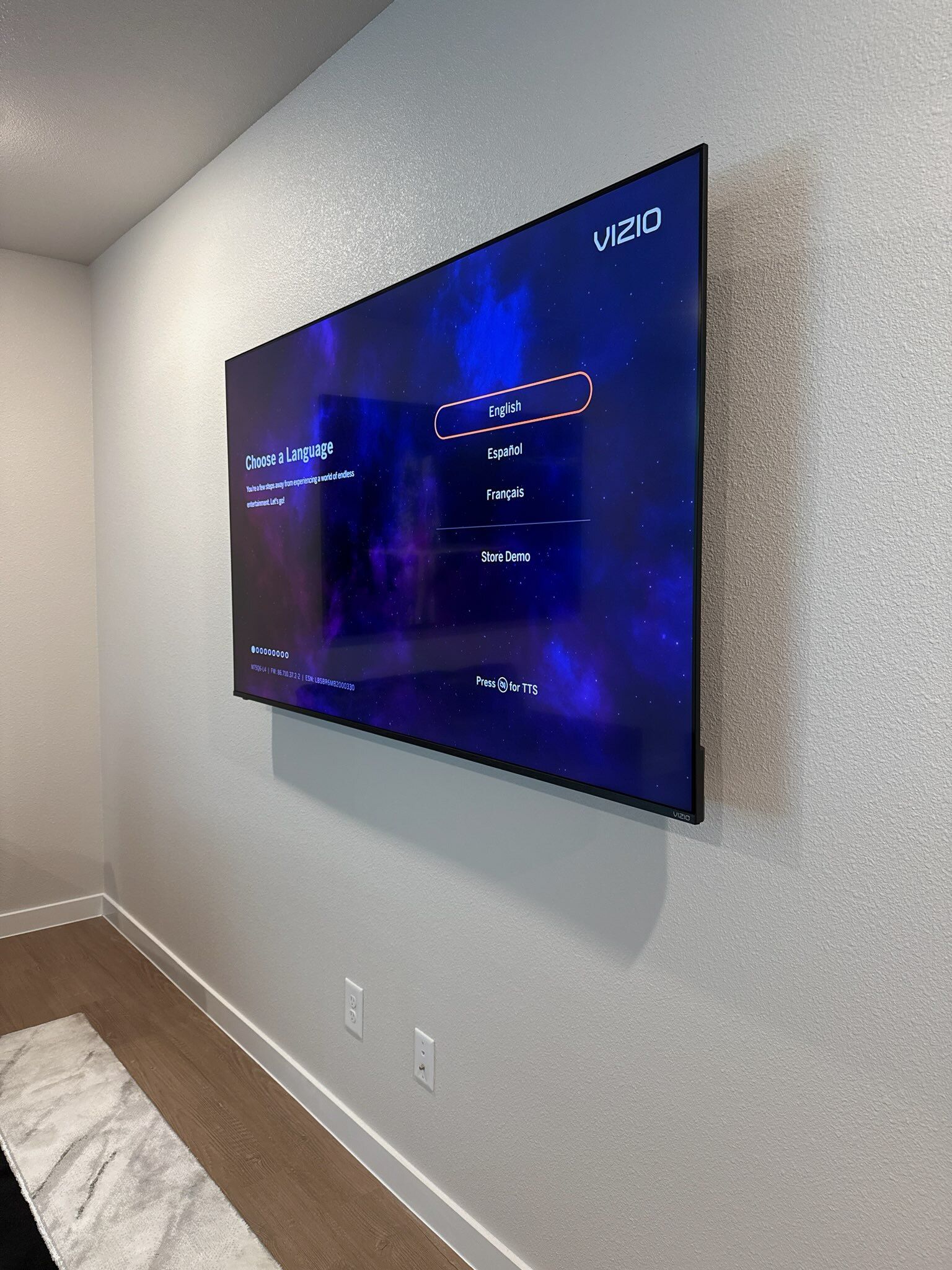 A large, mounted TV on a textured gray wall, casting a shadow. A rug and flooring are visible below.