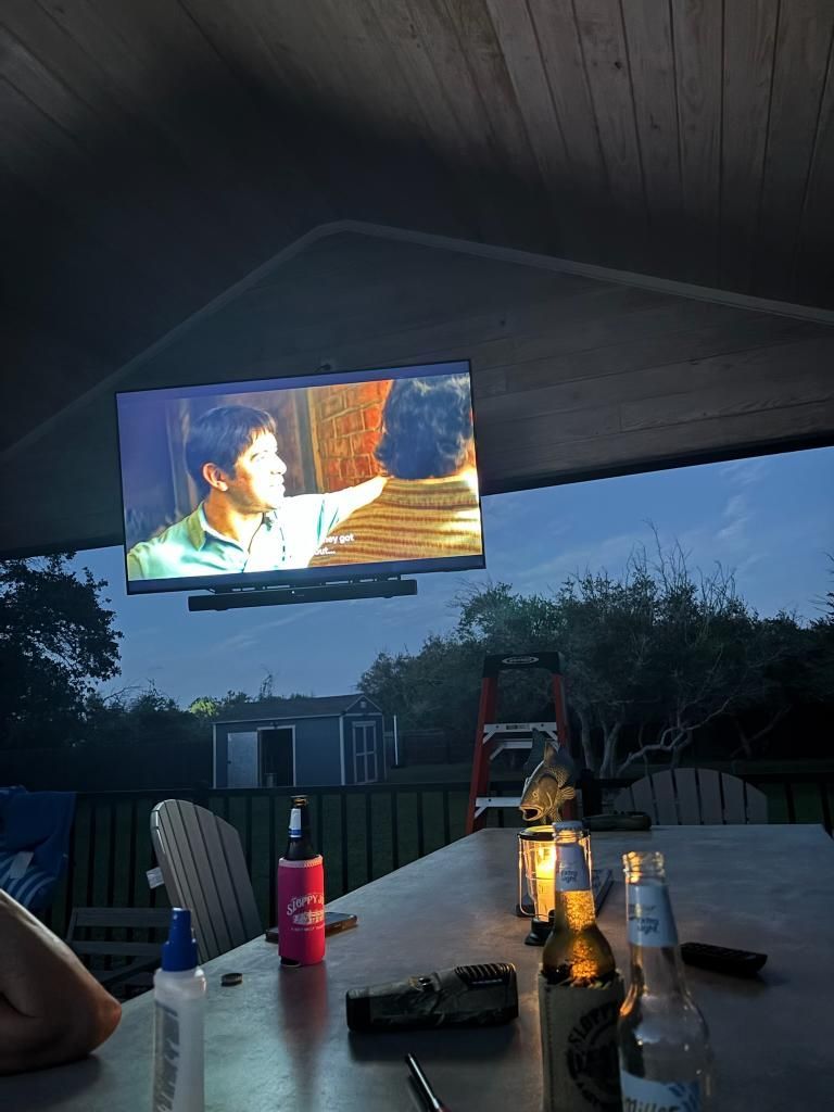 TV screen showing two men; outdoor bar setup with drinks, a small shed, and trees at dusk.