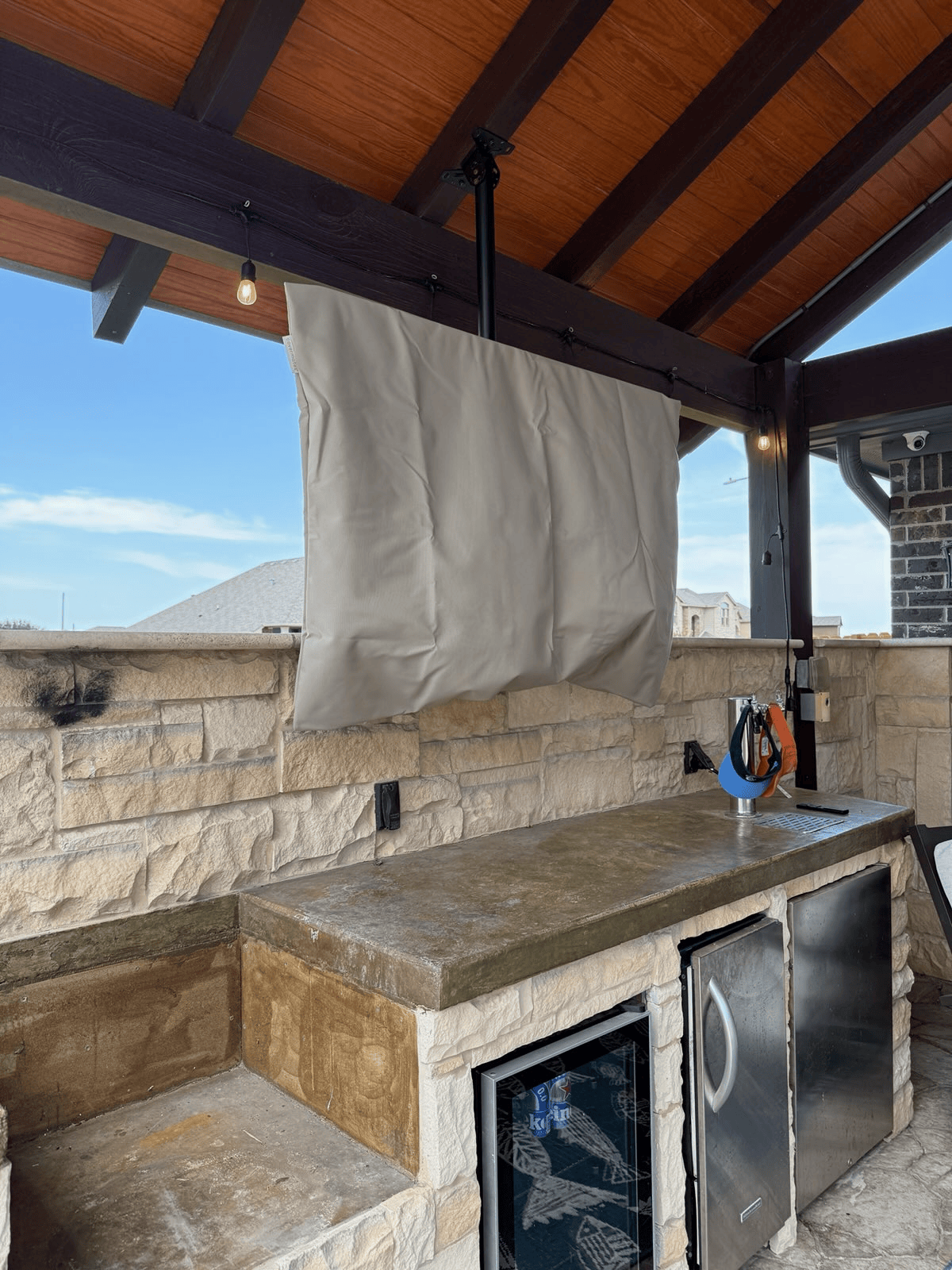 Outdoor kitchen with a TV covered by a tan fabric. Counter, cabinets, and stone wall visible.