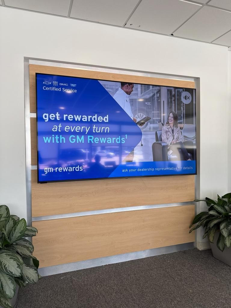 Large display promoting GM Rewards, mounted on a wood-paneled wall with plants.