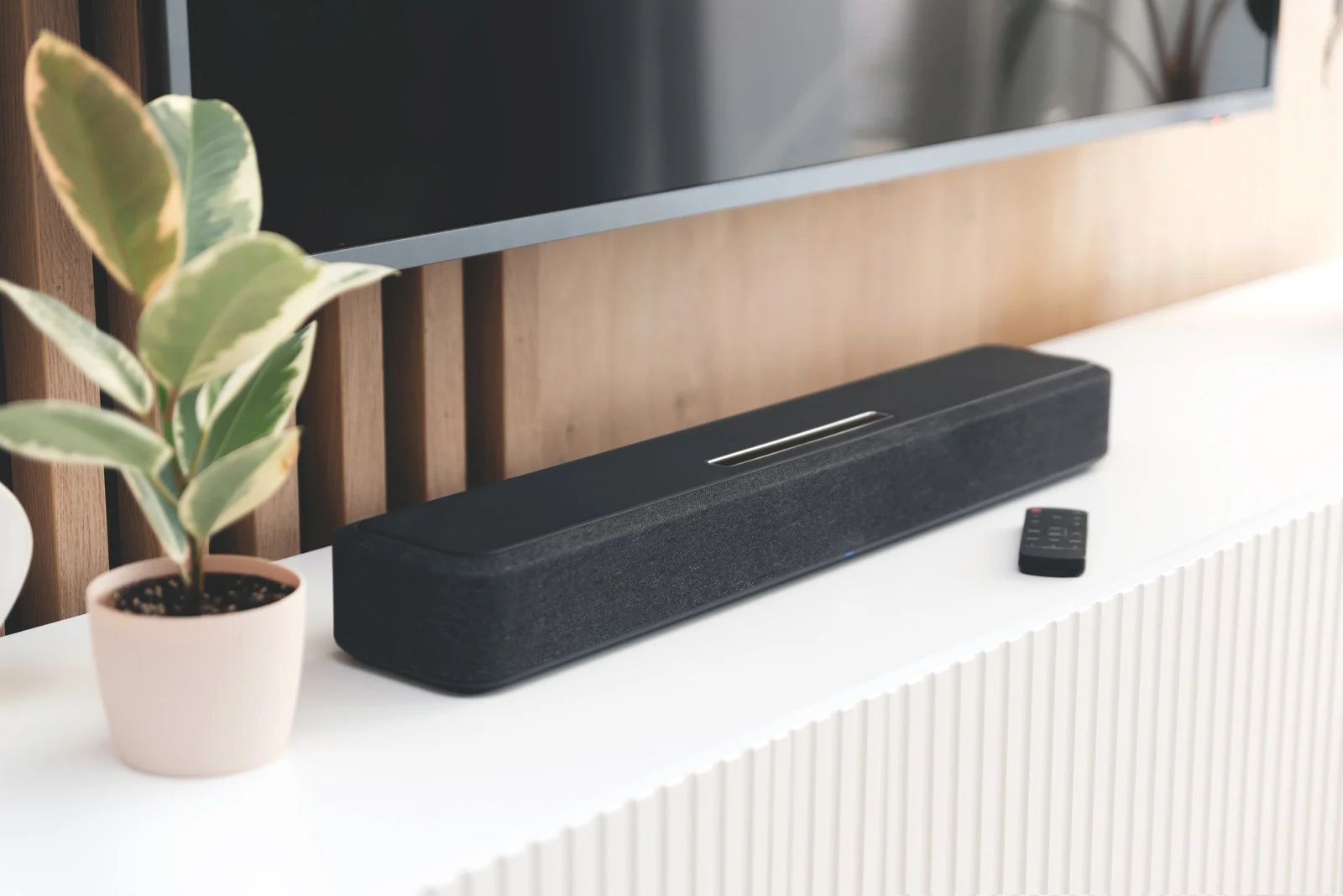 Black soundbar with remote on white cabinet; plant and TV in background.