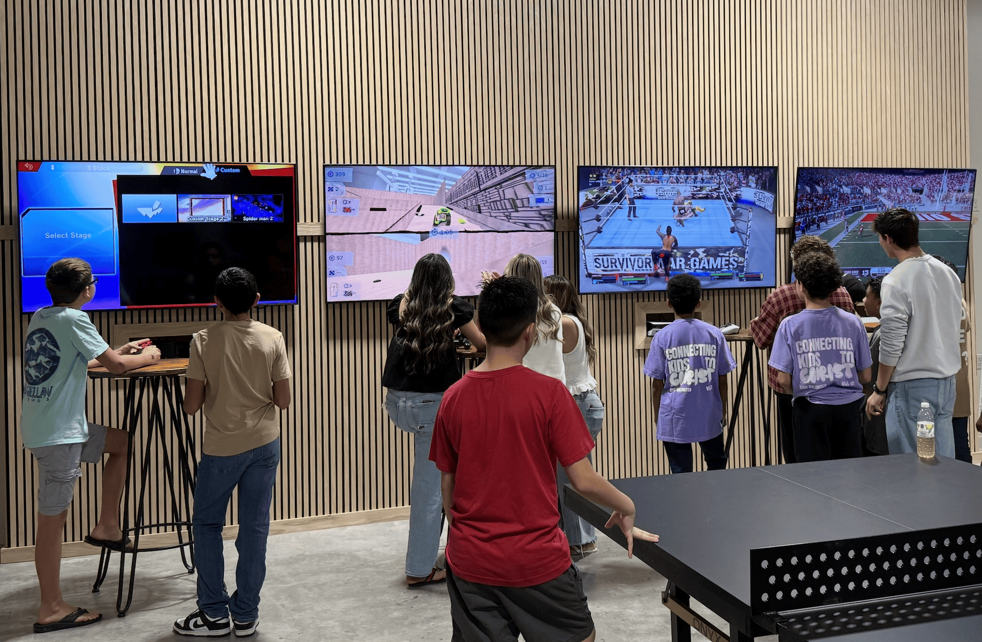 Group of people watching multiple TVs, some playing games, in a room with a ping pong table.