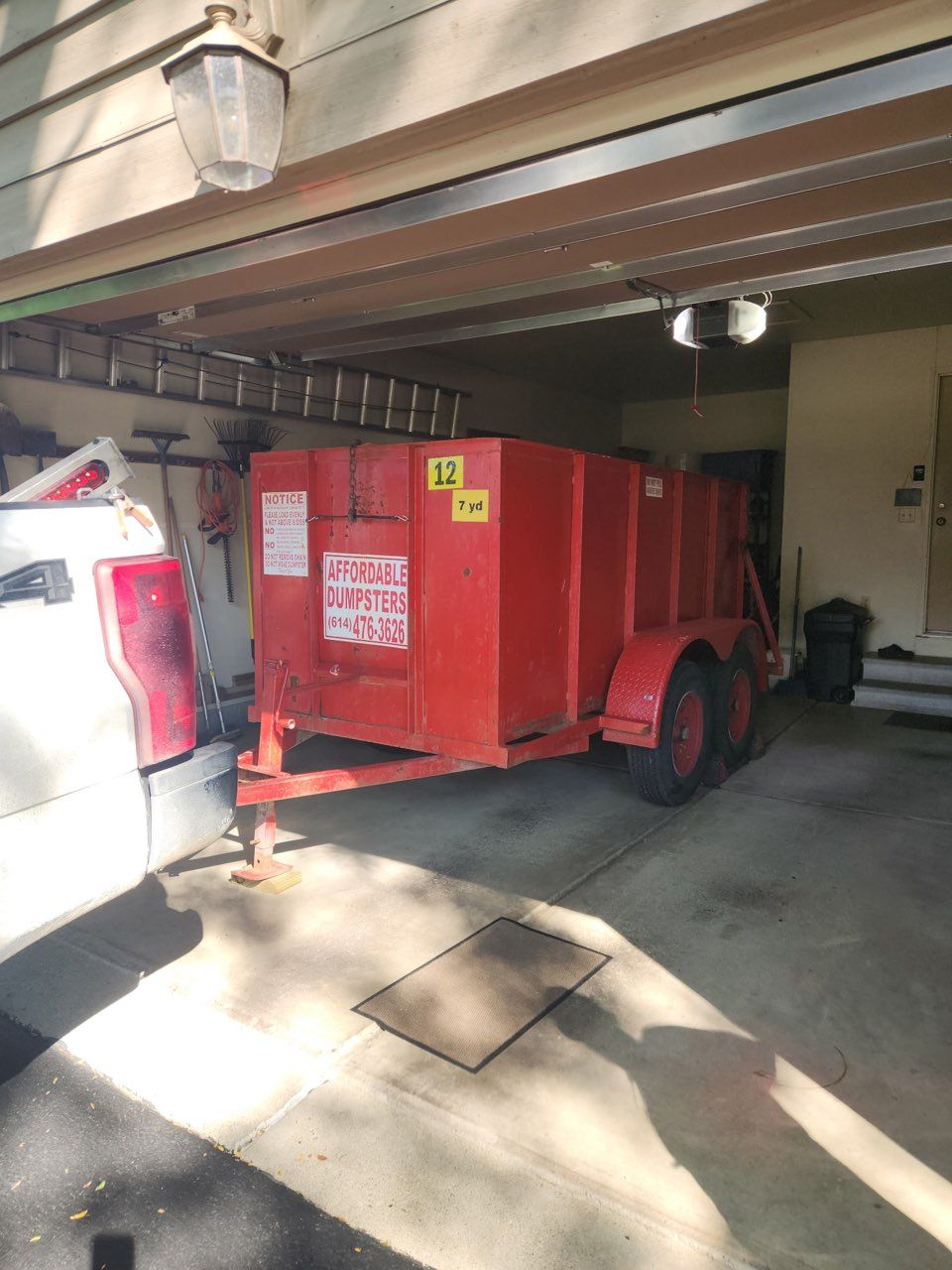 Red utility trailer parked under a carport beside a white truck and garage door