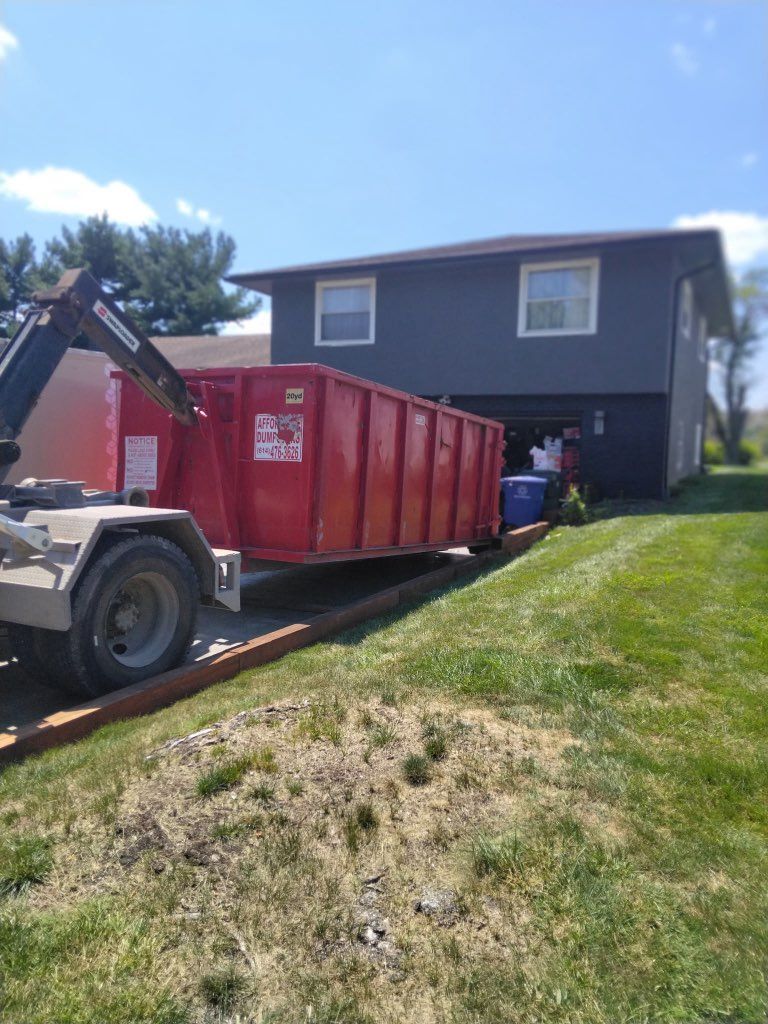 Worker loading a red dumpster onto a truck beside a dark house on a sunny slope