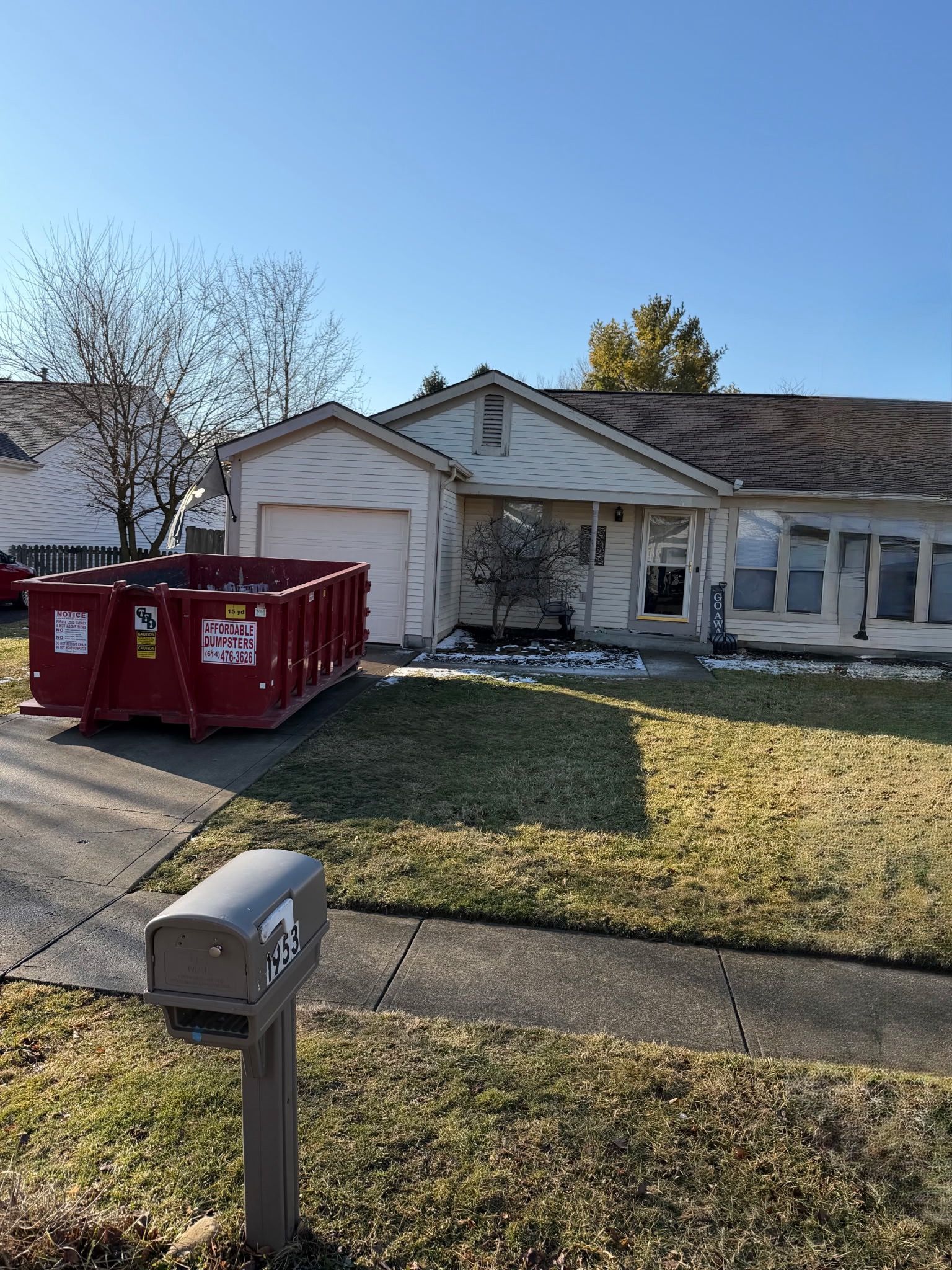 A one-story home with light siding and a driveway, featuring a large red dumpster parked in front on a sunny day.