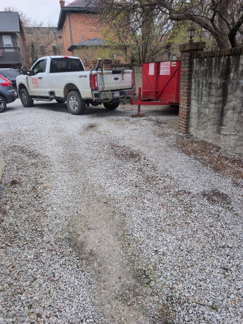 A garbage truck is parked in front of a building, carrying a large garbage bin in its truck bed. A white pickup truck is parked on a gravel driveway, hitched to a red metal dump trailer near a brick wall.