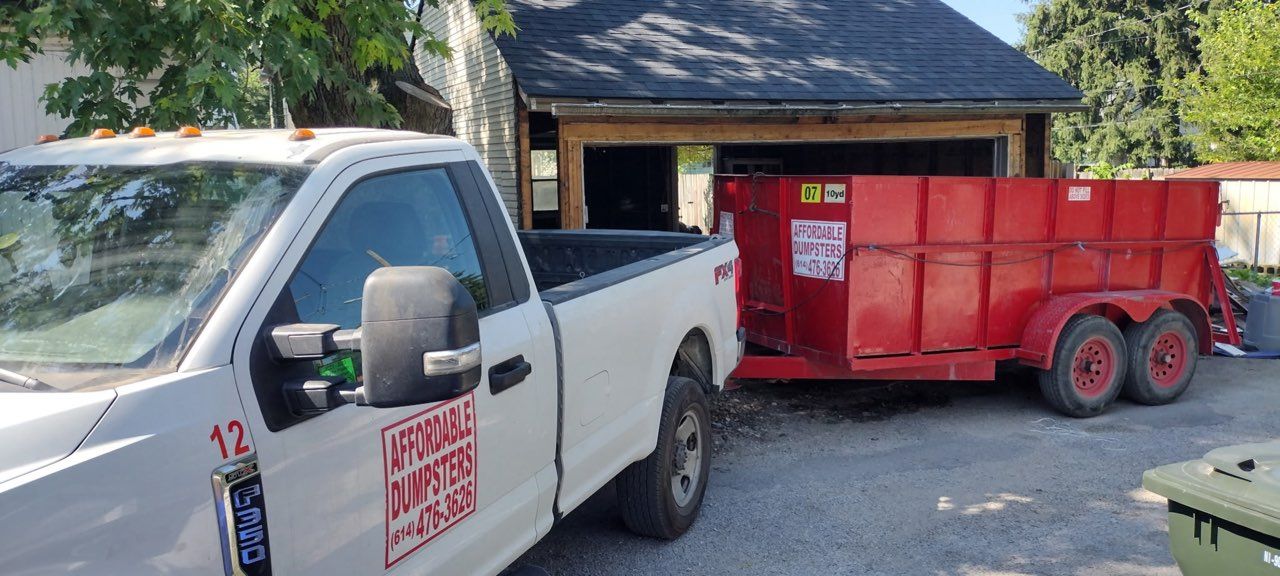 White pickup truck towing a red dump trailer beside a building in a driveway