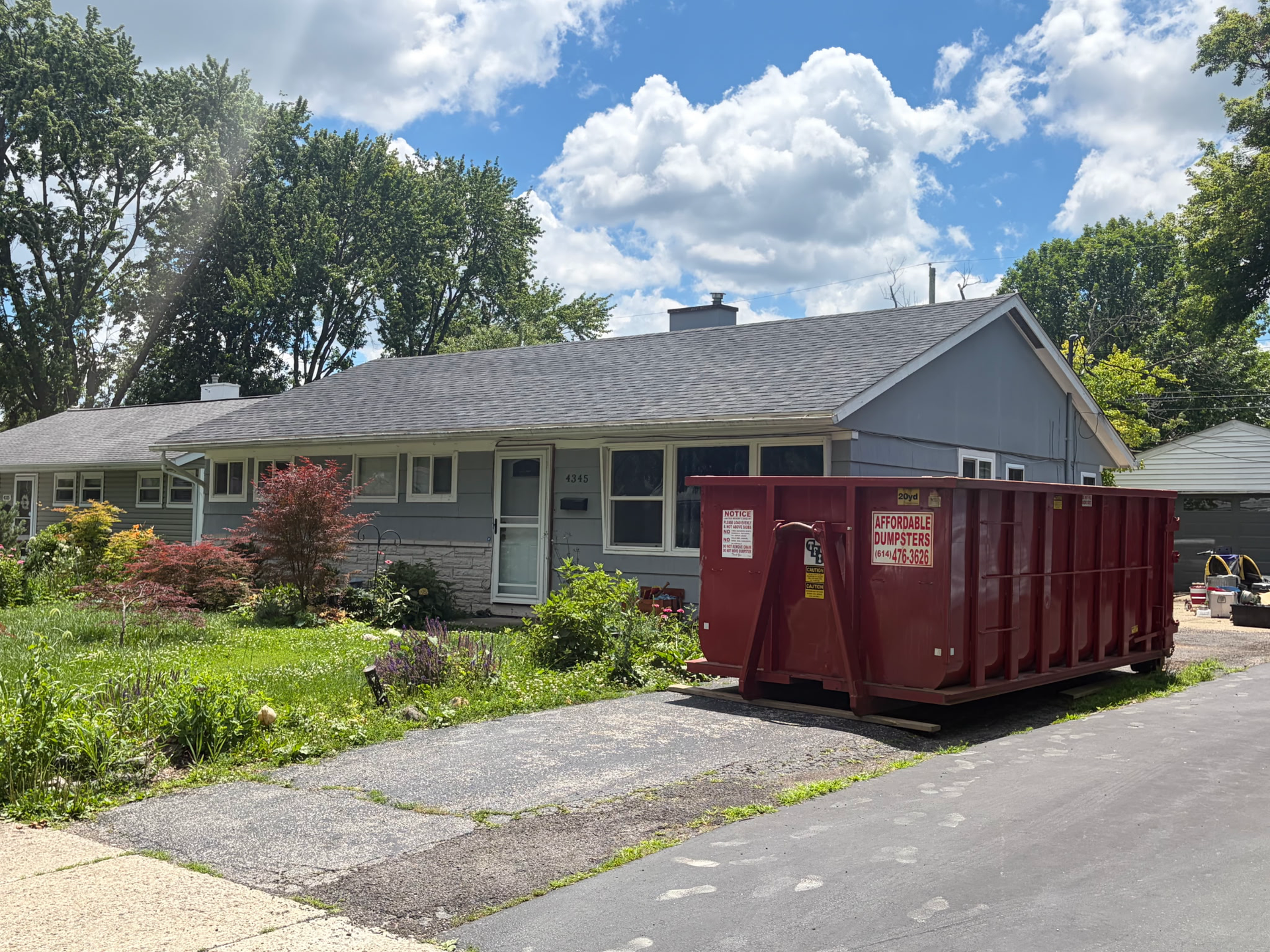 Gray house with a red dumpster in the driveway, surrounded by greenery under a cloudy sky