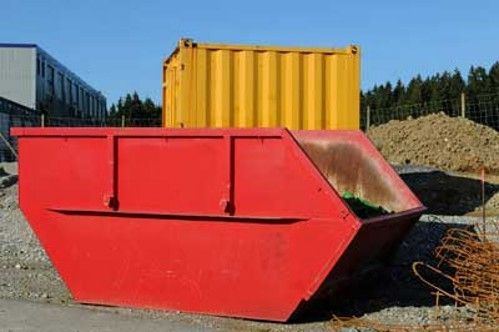Large red industrial dumpster with a yellow container on top in a gravel yard