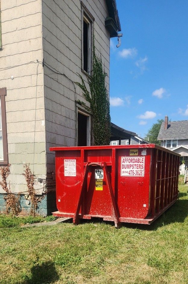 Red construction dumpster beside a house with ivy, on a grassy yard under a blue sky Red construction dumpster beside a house with ivy, on a grassy yard under a blue sky