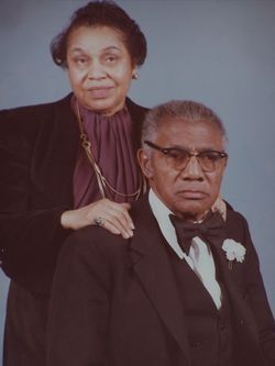 Formal portrait of an elderly couple in black attire, the woman standing behind the seated man on a gray background