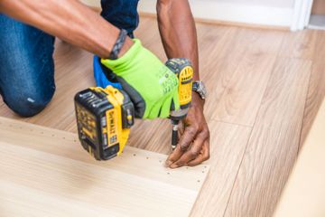 Person installing flooring with a cordless drill, wearing green gloves on a wood floor
