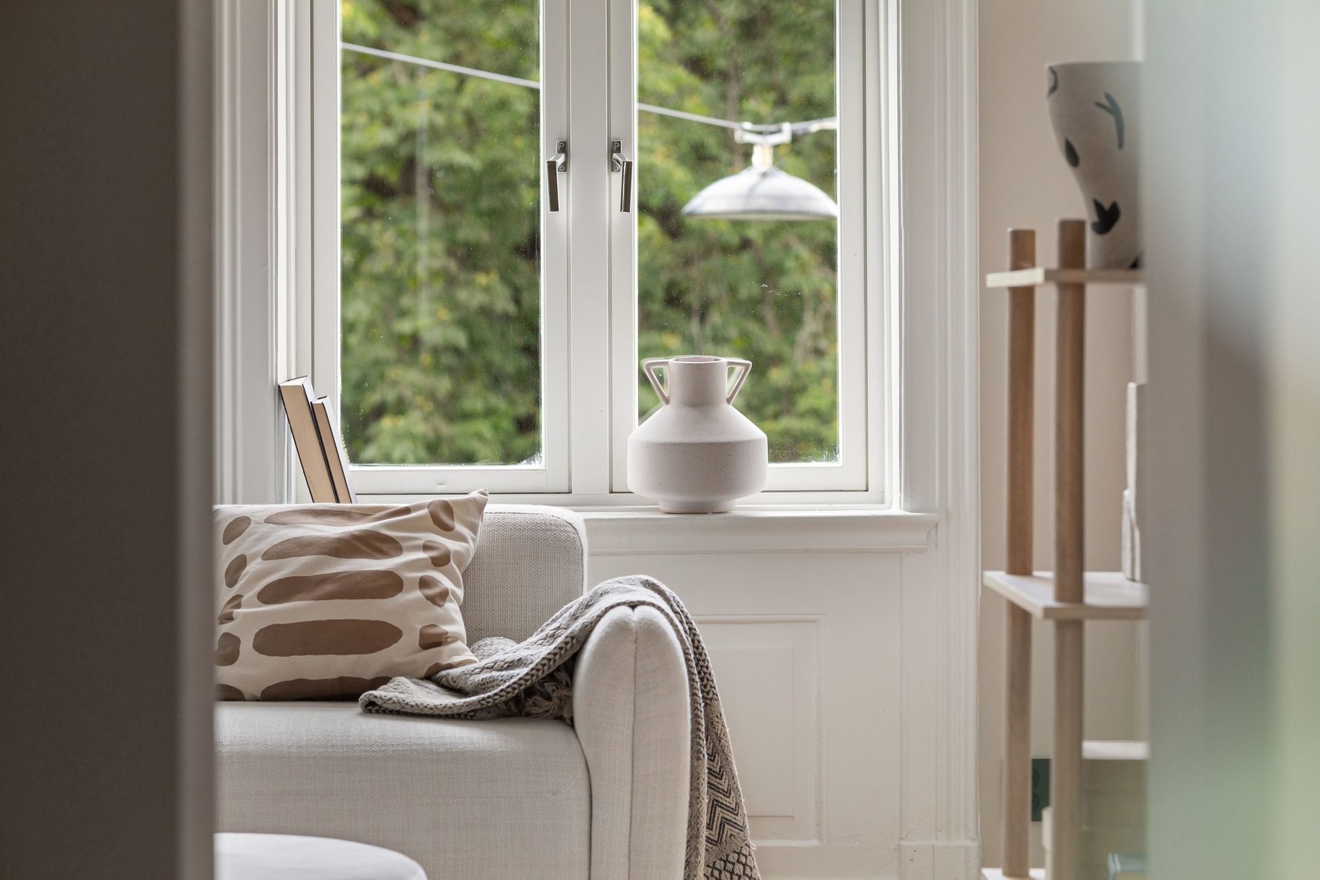 Cozy beige chair by a bright window with pillows, a draped throw, and a small vase on the sill
