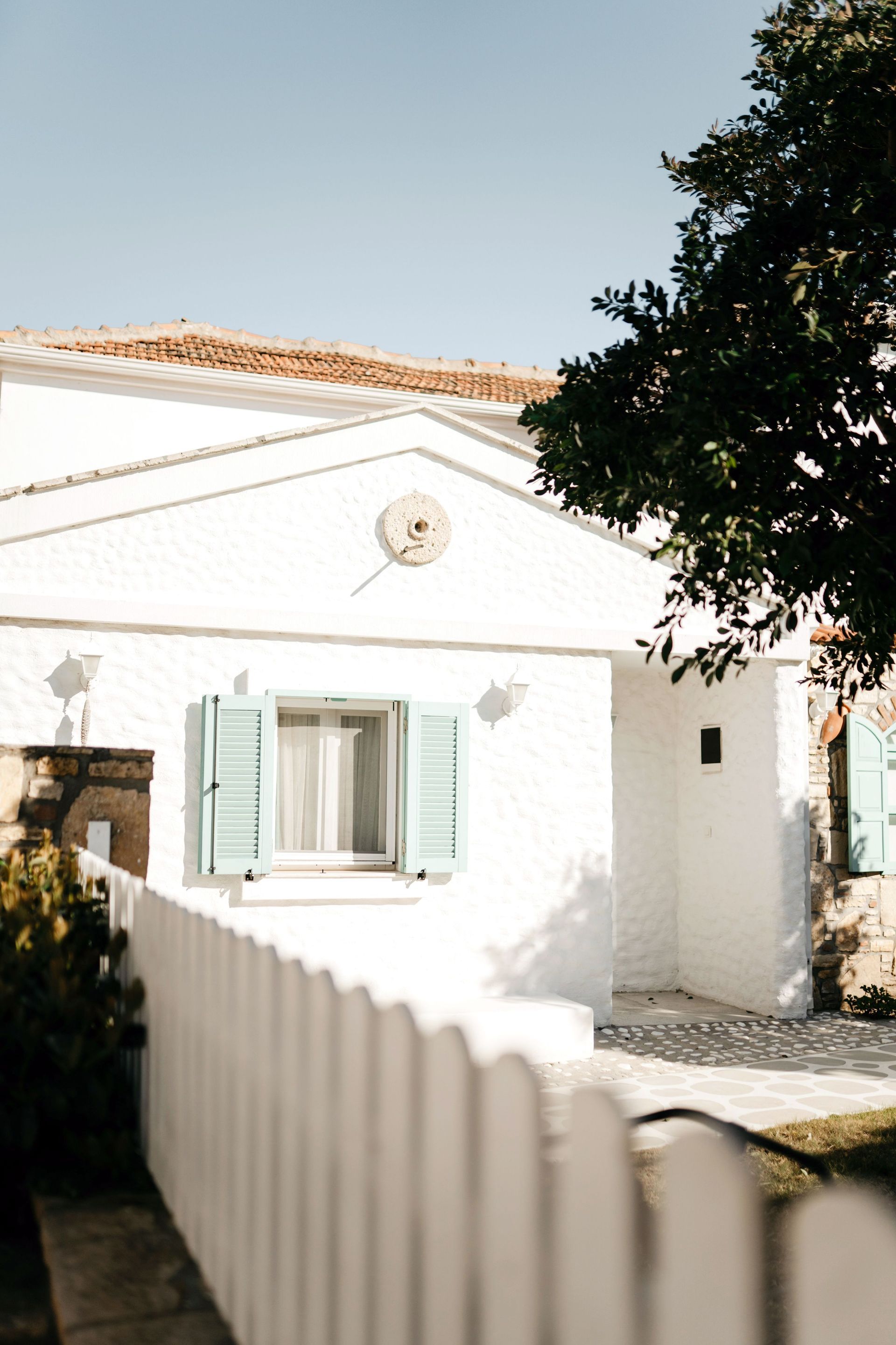 White house with mint shutters, sunlit courtyard, and a tree casting shade in the foreground