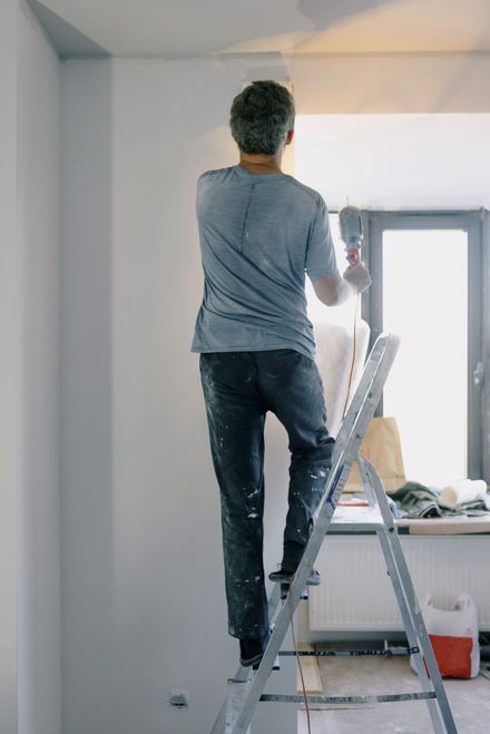 Person standing on a ladder painting a white wall near a window.