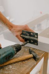 Hands using a drill to attach a metal hinge to a white wooden board on a workbench.