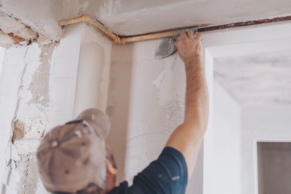 Person repairs a ceiling corner, applying plaster to cracks around exposed pipes.