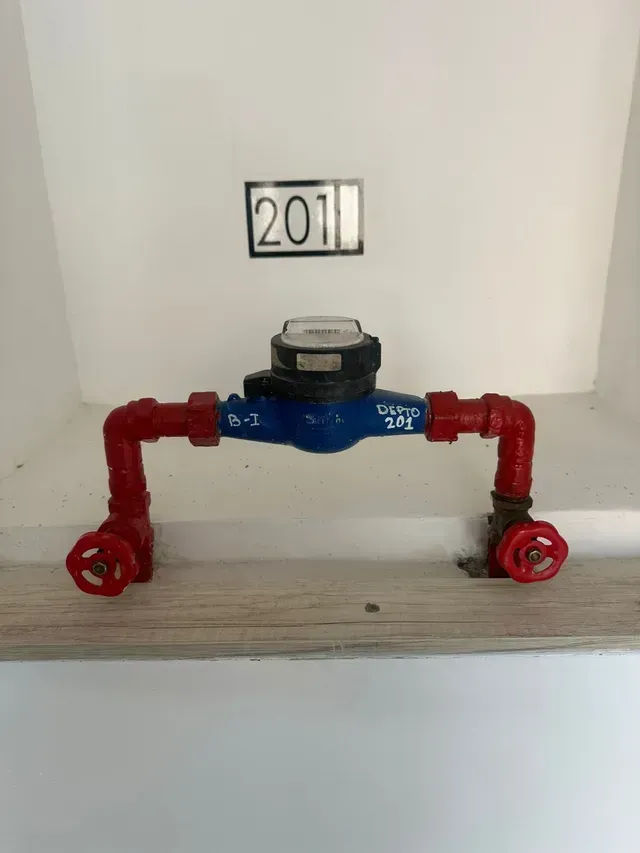 Contador de agua azul con tuberías y válvulas rojas montado en una pared, con la etiqueta “2011” en la parte superior.