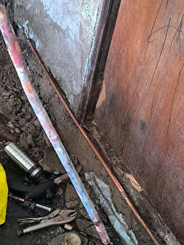 Esquina de pared mohosa con tuberías pintadas inclinadas, revestimiento de madera y herramientas de construcción en el suelo.