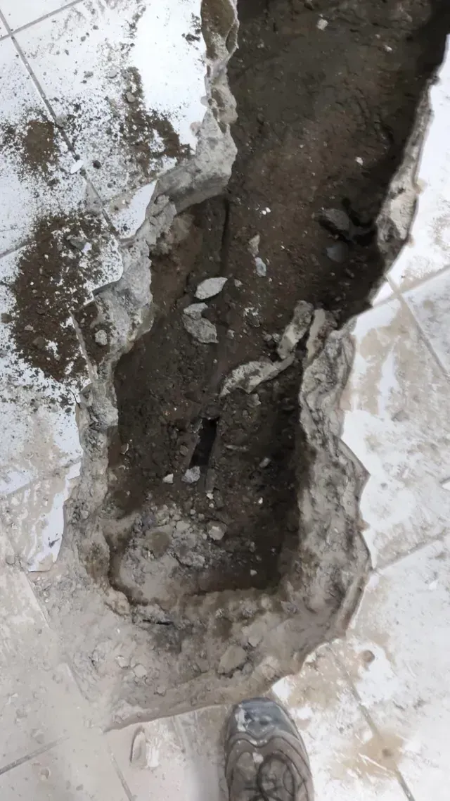 Primer plano de un agujero de hormigón rugoso lleno de tierra y escombros sobre un suelo blanco dañado.
