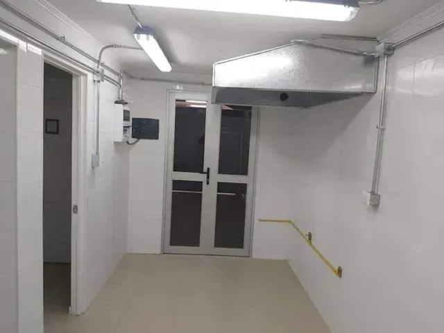 Pasillo blanco con luces fluorescentes, puertas dobles de cristal y una barandilla amarilla en la pared derecha.